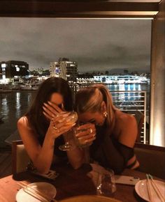 two women sitting at a table with wine glasses in front of their faces and the city lights behind them