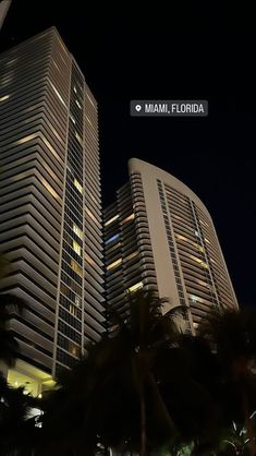 some very tall buildings at night time with palm trees in the foreground and lights on