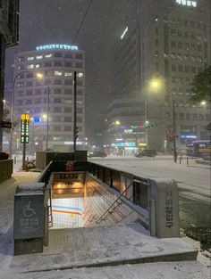 a city street at night with snow falling on the ground and buildings in the background