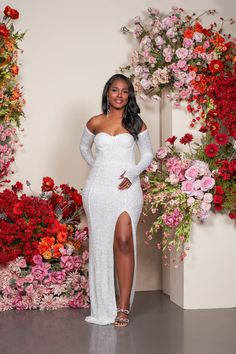 a woman standing in front of flowers wearing a white gown with long sleeves and thigh high slit