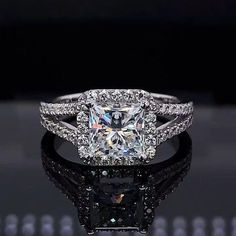 an engagement ring with a princess cut diamond surrounded by round brilliant pave diamonds on a black reflective surface