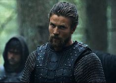 a man with a beard and armor standing next to another man in the woods,