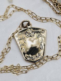 This is a "once in a lifetime" piece of estate jewelry.  This would make a lovely, thoughtful gift for your sweetheart. This very special antique locket necklace is made of highly polished solid yellow gold.  The locket is 10K and the chain is 14K.  Both are stamped accordingly. The locket has a unique shield shape which has a beautiful floral forget-me-not design on the front and a monogram on the back.  The locket is a little over one inch long.  The inside still holds the original black and white portrait of a handsome gentleman.  The photo is held in place by the original frame and cover.  I imagine there is a wonderful love story attached to this piece.  The necklace is a mini paperclip style chain with a spring ring clasp.  The clasp is stamped 585 (14K).  The chain is 18 inches long Brass Heart Pendant Wedding Jewelry, Brass Heart Pendant Jewelry For Wedding, Brass Heart Pendant Jewelry For Anniversary, Elegant Brass Jewelry For Valentine's Day, Elegant Brass Necklaces For Valentine's Day, White Gold Vintage Charm Pendant Jewelry, Vintage White Gold Pendant Jewelry, Heirloom 14k Gold Heart Jewelry, Vintage Charm Yellow Gold Heart Pendant Jewelry