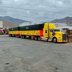 a large yellow semi truck parked in a lot next to other trucks and buildings with mountains in the background