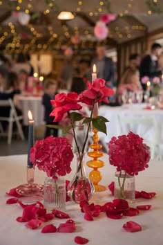 Round table decor in a barn venue. Round table with a white tablecloth. Pink and orange contemporary glass candlesticks with coloured candles. Single stem pink hydrangeas and pink roses in bud vases. Pink rose petals are scattered around. Single Vase Centerpiece Wedding Ideas, Pink And Orange Bud Vases, Single Stem Table Decor, Pink And Orange Wedding Centerpieces, Hydrangea Bud Vase, Hot Pink And Orange Party Decorations, Single Stem Centerpiece, Roses In Bud Vases, Pink Wedding Table Settings