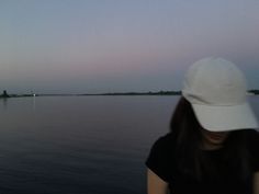 a woman wearing a white hat standing in front of a body of water at dusk