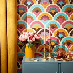 a vase with flowers and two candles on a side table in front of a colorful wallpaper