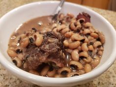 a white bowl filled with beans and meat on top of a marble counter next to a silver spoon