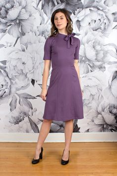 "This demure dress will have you walking comfortably into any meeting or boardroom with an elegant, feminine stride.  Pullover style with a mock neck neckline and bow detail, short sleeves ideal for layering under a jacket, and a super flatting fit and flare silhouette. Heavy weight double knit ponte fabric is comfortable, wrinkle resistant and easy to care for.  Composition: 65% Rayon, 30% Nylon, 5% Spandex Care: Machine wash cool, hang to dry, medium iron or dry clean. Garment Measurements: La Fitted Purple Midi Dress For Office, Purple Fitted Midi Dress For Office, Elegant Purple Office Dress, Purple Knee-length Office Dress, Purple Knee-length Dress For Office, Purple A-line Dress For Workwear, Purple Fitted A-line Midi Dress, Elegant Purple Midi Dress For Office, Chic Purple Dress For Office