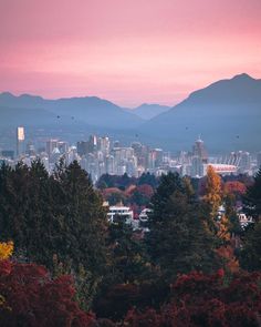 the city is surrounded by mountains and trees with colorful foliage in front of it at sunset