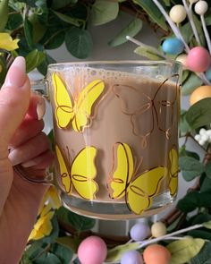 a person holding up a glass mug with yellow butterflies painted on it and surrounded by flowers