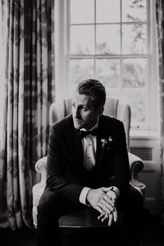 a man in a tuxedo sitting next to a window with curtains on it