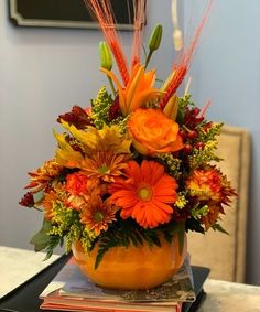 a vase filled with orange flowers and greenery on top of a table next to a stack of books