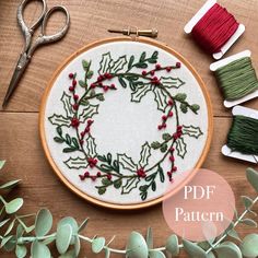 a cross stitch wreath with red berries and green sprigs is surrounded by scissors