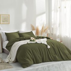 a bed with green sheets and pillows in a white room next to a window filled with plants