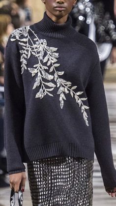 a model walks down the runway in a black sweater and silver skirt with white flowers on it