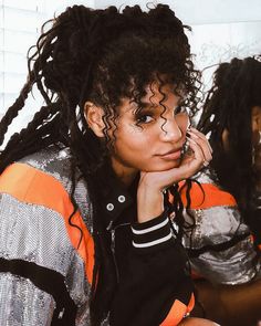 a woman with dreadlocks sitting in front of a mirror wearing an orange and black jacket