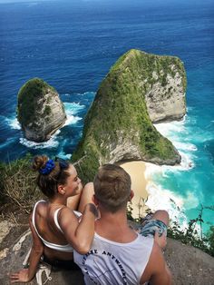 two people sitting on the edge of a cliff looking at the ocean