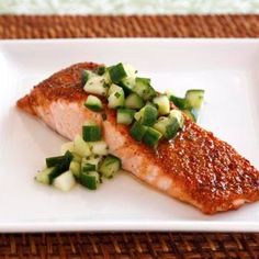 a piece of fish with cucumbers and sauce on a white plate sitting on a wicker place mat