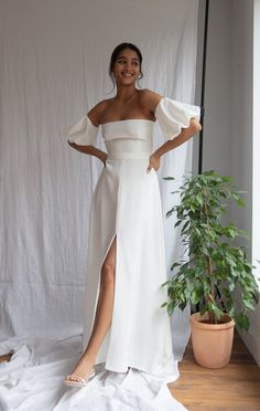 a woman standing in front of a white backdrop wearing a dress with an off the shoulder design