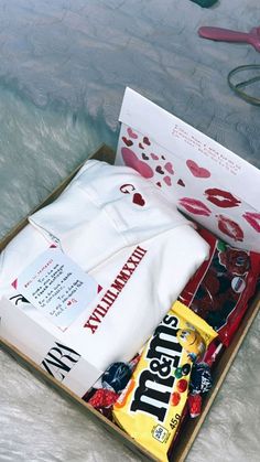 an open box containing candy, t - shirts and scissors on a bed with white fur