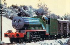 a thomas the tank engine is coming down the tracks in the snow with trees and bushes behind it