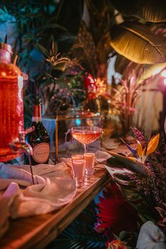 This shows a close up of a wine glass with 3 shot glasses on a bamboo bench. The scene is dramatically lit by white back lighting. In front of the bench is a large flower arrangement with tropical flowers. Next to the glassware is a white table runner and some faux plants in the background. Drinks Area, Grey Photography, Tropical Florals