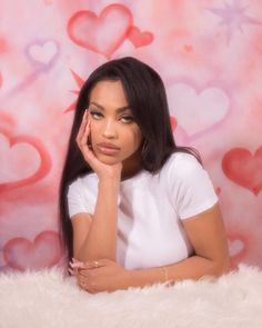 a woman sitting in front of a pink wall with hearts on it and her hand under her chin