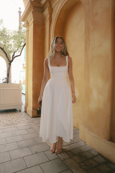 a woman in a white dress is standing by a wall and smiling at the camera