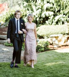 a man and woman in formal wear walking through the grass with trees in the background