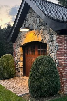 a small brick house with a black roof and two bushes in front of the door