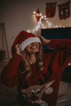 a woman wearing a christmas sweater and santa claus hat smiles while sitting on her bed