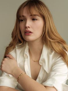 a woman with long red hair wearing a white shirt and gold bracelet sitting in front of a gray background