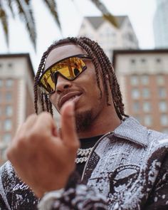 a man with dreadlocks and sunglasses pointing to the side while standing in front of some buildings