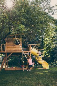 two children playing on a wooden play set in the grass under a tree with a yellow slide