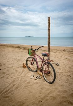 a pink bike parked on top of a sandy beach next to a wooden pole and the ocean