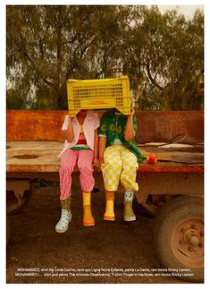 two people sitting on the back of a truck with a basket over their head and fruit in front of them