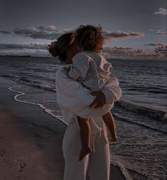 a woman holding a child on the beach at sunset with clouds in the sky behind her