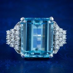 A magnificent vintage cocktail ring made in the Art Deco fashion with a colossal emerald cut aquamarine in the centre, weighing approx. 15ct with a pleasing, deep ocean blue hue.  The aqua is complemented by an audience of twenty-four sparkling brilliant cut diamonds arranged in a graduated, stepped design down each shoulder. They're 0.05ct each (approx. 1.20ct total) and have superb SI1 clarity - H colour.  The robust gallery is modelled entirely in 18ct white gold and displays the stones high Formal Light Blue Diamond Ring, Formal Aquamarine Diamond Ring With Brilliant Cut, Aquamarine Brilliant Cut Diamond Ring For Formal Occasions, Classic Blue Radiant Cut Emerald Ring, Modern Blue Baguette Cut Emerald Ring, Luxury Gia-certified Emerald Cut Topaz Ring, Formal Light Blue Topaz Ring, Blue Radiant-cut Emerald Ring In Fine Jewelry Style, Formal Aquamarine Diamond Ring With Prong Setting