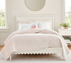 a white bed with pink comforter and pillows in a light colored room next to two windows