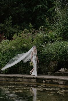 a woman in a wedding dress is walking by the water with her veil flying over her head