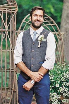 a man wearing a vest and tie standing in front of a wooden trellis with flowers