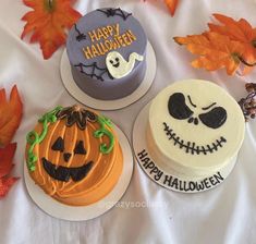 three decorated halloween cakes sitting on top of a white sheet covered table with leaves and pumpkins