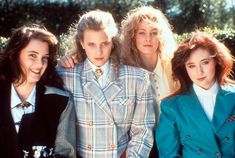 four women are posing for a picture together in front of trees and bushes, one is wearing a plaid jacket