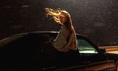 a woman standing next to a black car in the rain with her hair blowing back