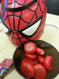 some red tomatoes in a bowl next to a spider - man costume helmet on a table