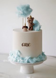 a one tiered cake with a teddy bear topper and clouds on the side