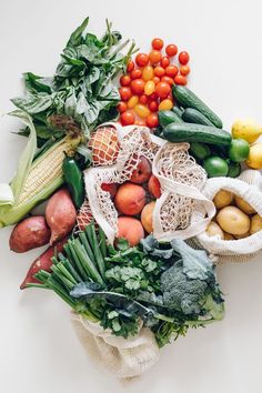 many different types of fruits and vegetables on a white surface with mesh bags over them