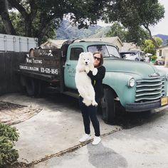 a woman standing next to an old truck holding a white dog in her arms and smiling at the camera