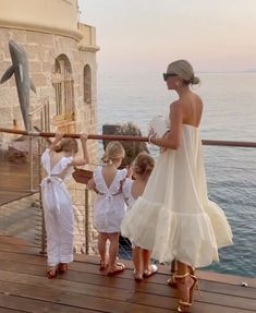 a woman and two small children on a pier looking at dolphins in the water behind them